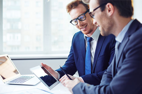 Two men wearing glasses, suits and ties holding an ipad and a cell phone smiling